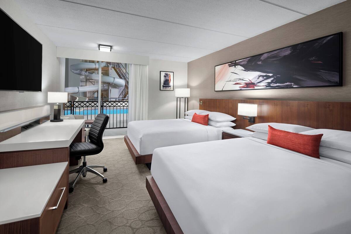 A hotel room with two beds, a desk, a chair, a TV, and a large window with a pool view.