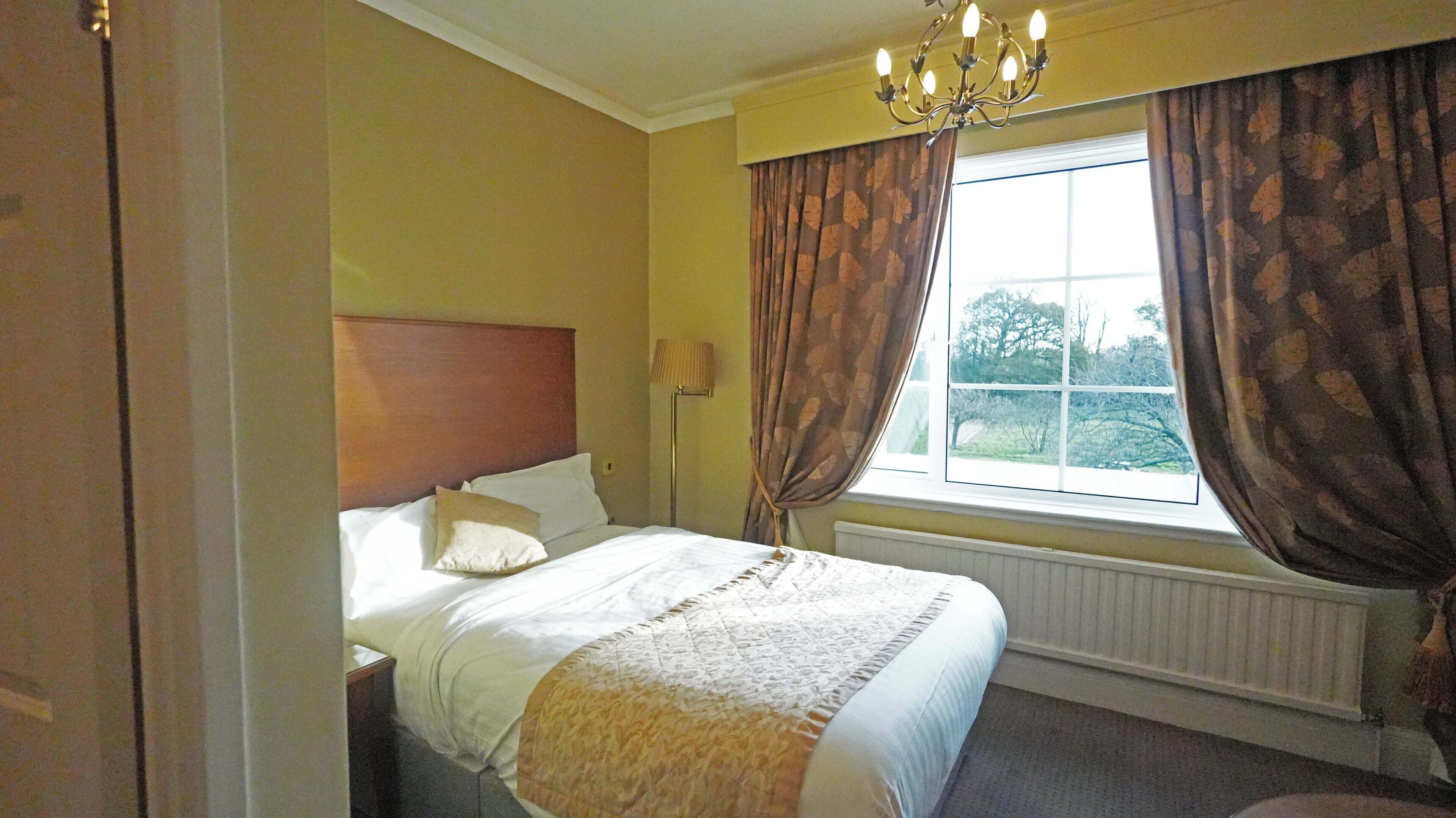 Una habitación de hotel con cama, mesita de noche, lámpara, ventana con cortinas y un candelabro.