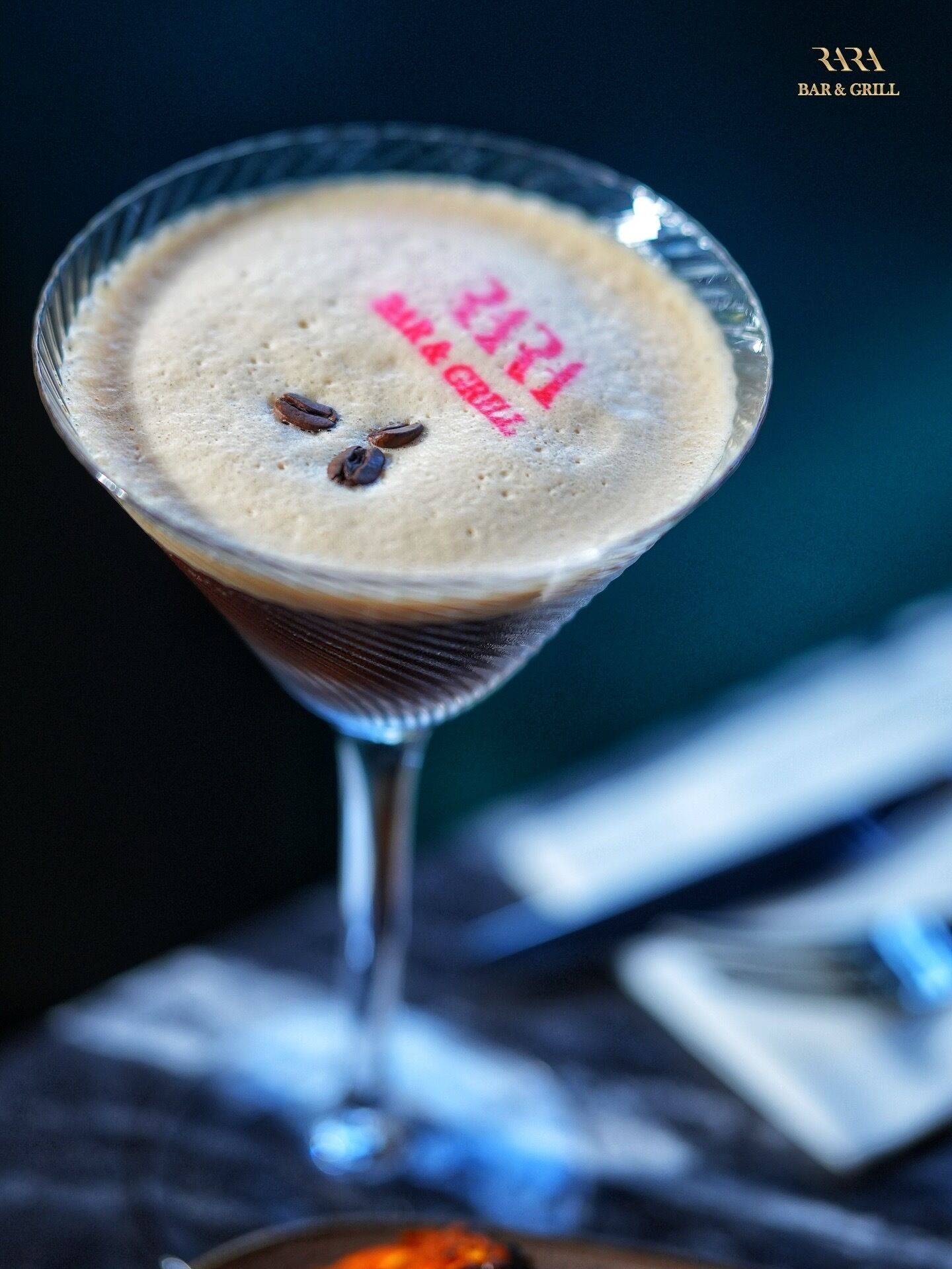 A martini glass with a frothy top, a dark background, and a napkin and a small orange object on the table.