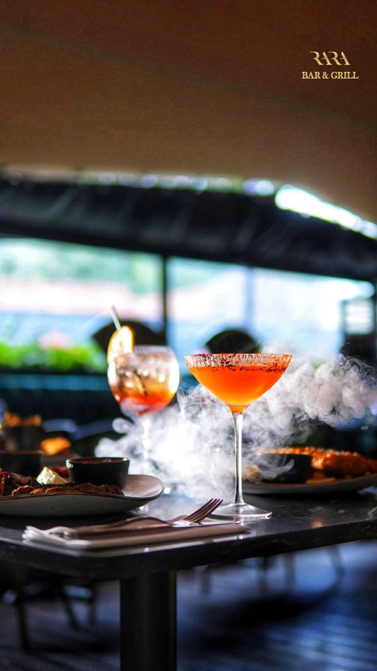 En barscene med to cocktails, en tallerken mad og udsigt til et bybillede.
