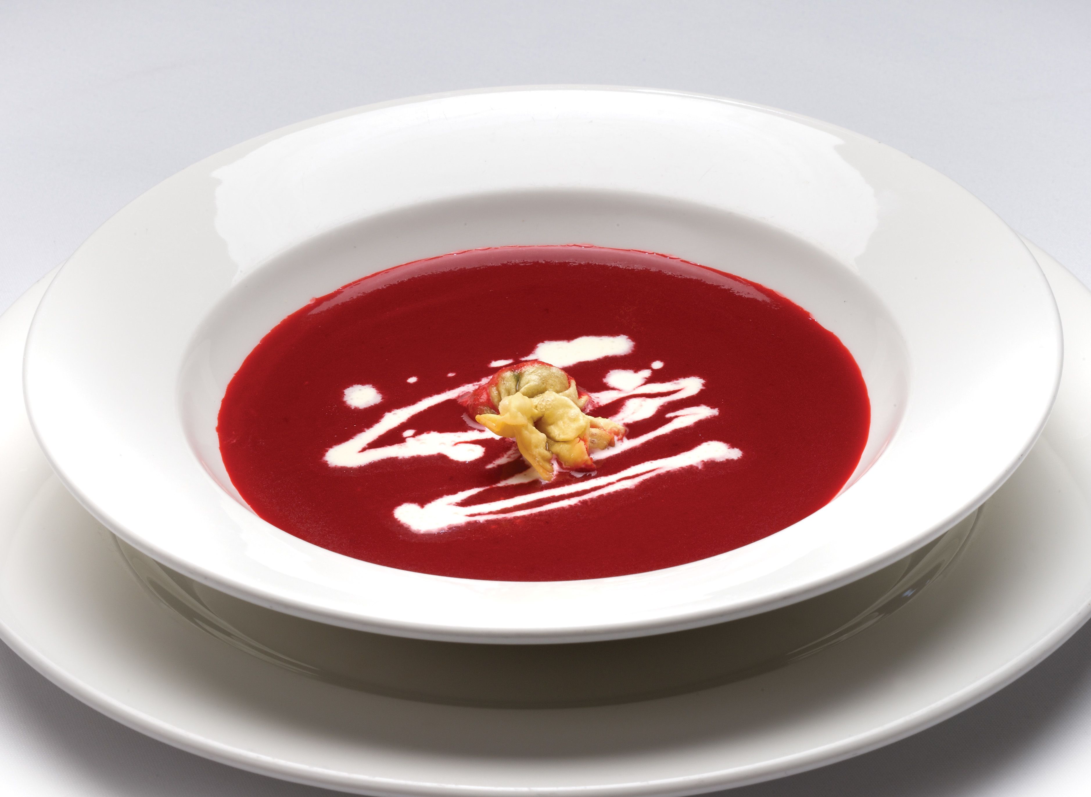 A white bowl of red soup with a dollop of cream and a piece of fruit on top, placed on a white plate.