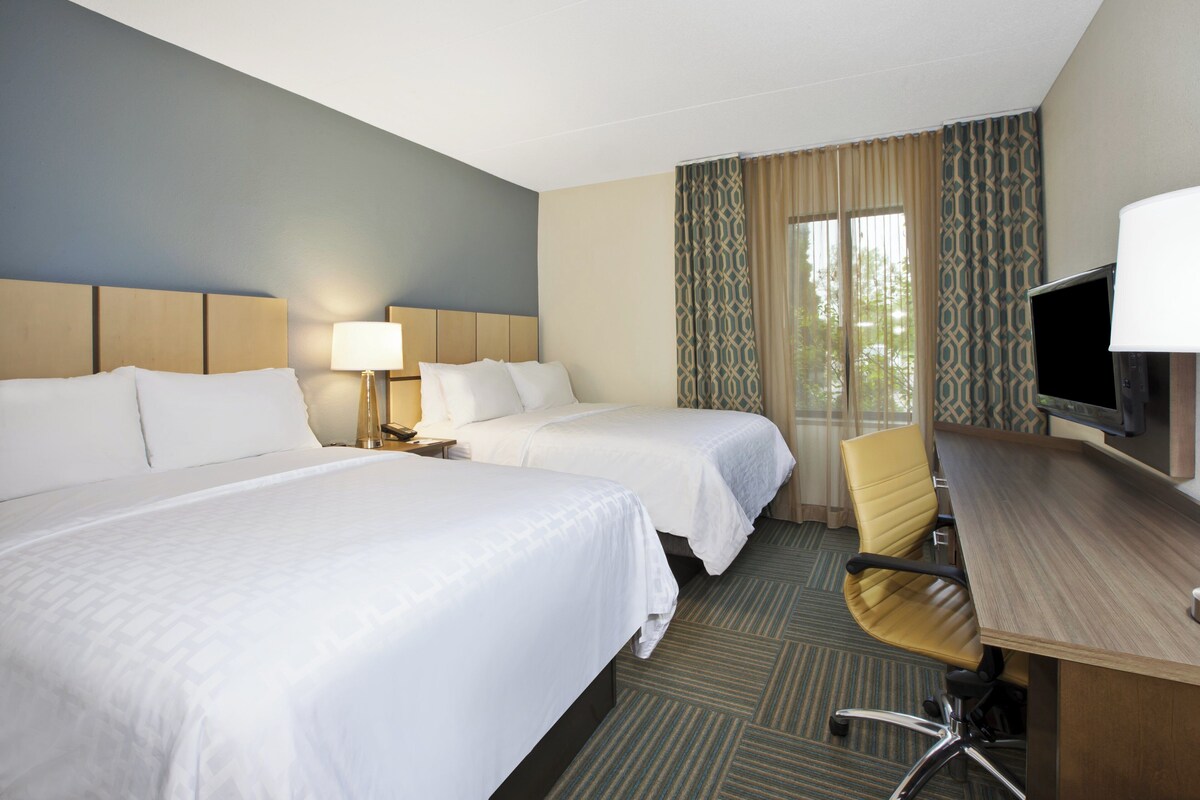A hotel room with two beds, a desk with a chair, a TV, and a window with curtains.