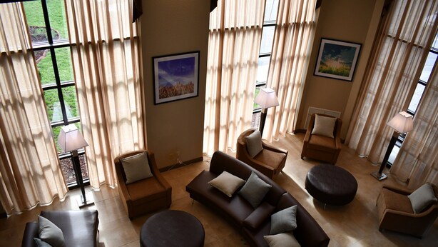 A spacious living room with a sectional sofa, round ottoman, and a small table. There are two lamps, a chair, and a view of the outdoors through large windows. - Candlewood Suites Grand Rapids Airport by IHG (Grand Rapids)