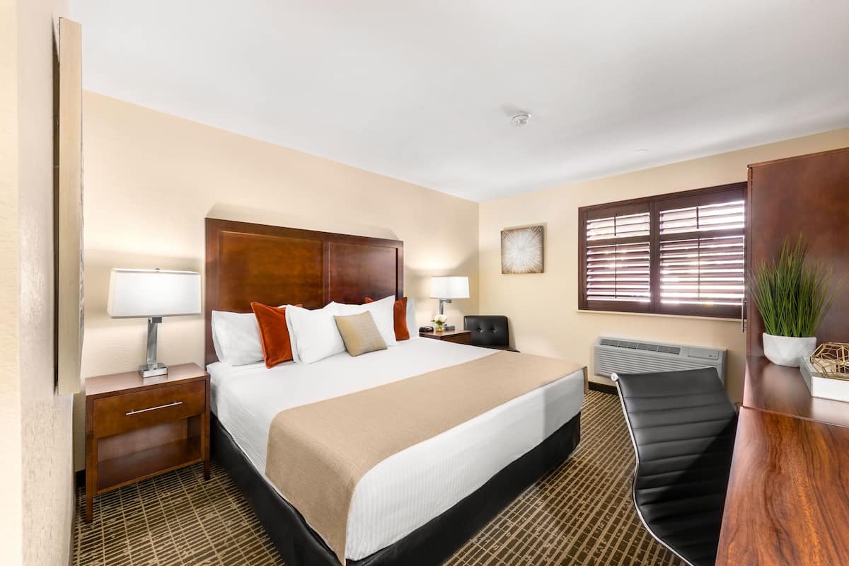 Habitación de hotel con una cama grande, mesitas de noche con lámparas, un escritorio con silla, un radiador y una ventana con persianas.