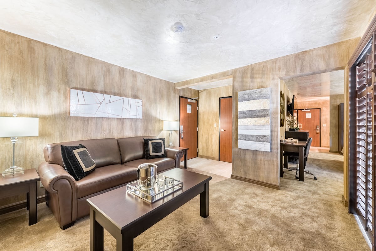 Una habitación de hotel moderna con un sofá de cuero, una mesa de centro con una bandeja, una lámpara y un cuadro abstracto grande en la pared.