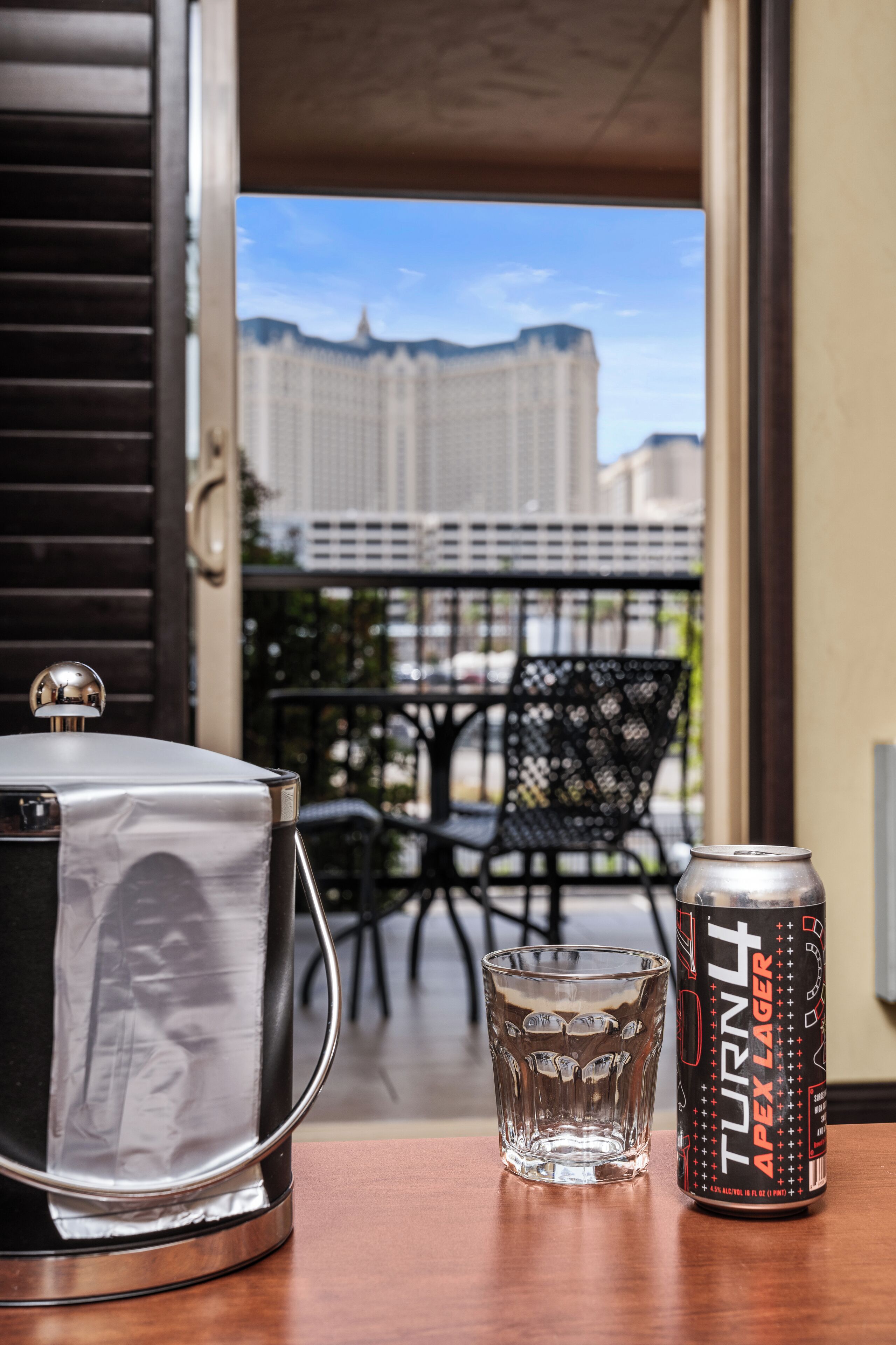 A table with a can of beer, a glass, and a kettle on a balcony with a view of buildings.