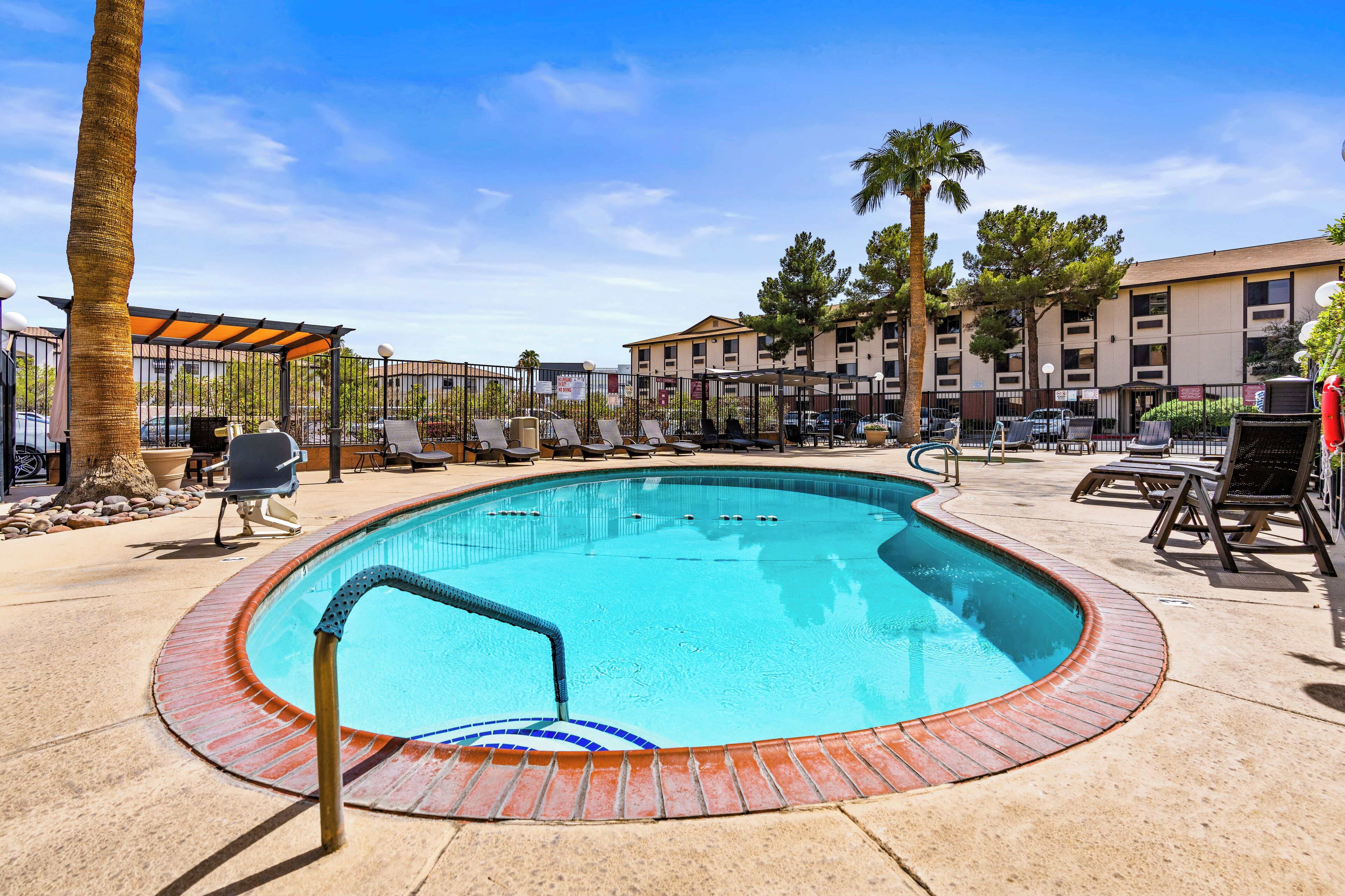 A swimming pool with lounge chairs and a covered seating area, surrounded by a fence and palm trees.