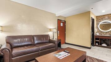 A hotel room with a brown leather sofa, a wooden coffee table, a small side table, a lamp, and a bathroom with a mirror and sink.