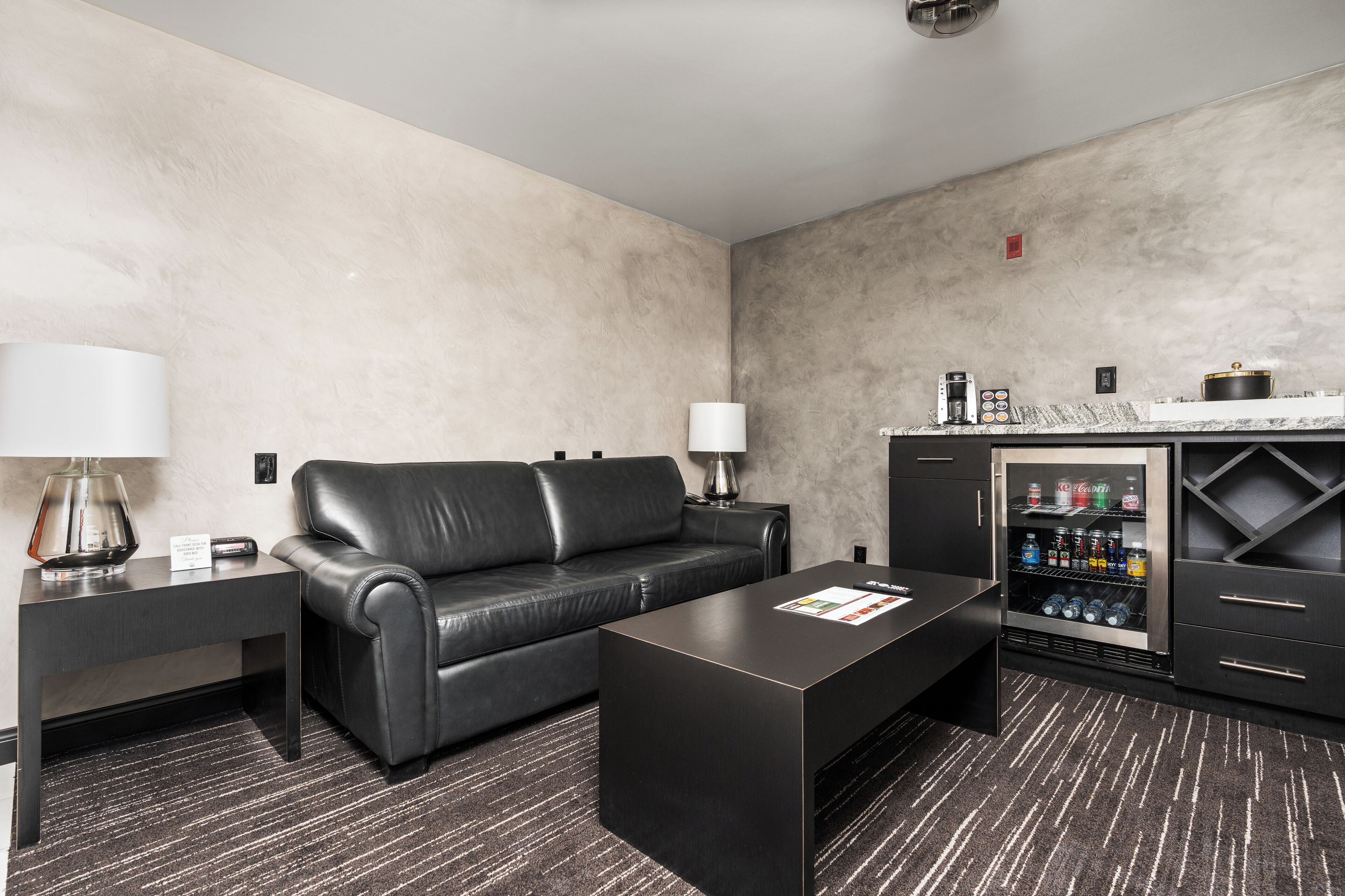 Ein modernes Hotelzimmer mit einer schwarzen Ledersofa, einem Couchtisch, einer Minibar und einem Schreibtisch mit Lampe.