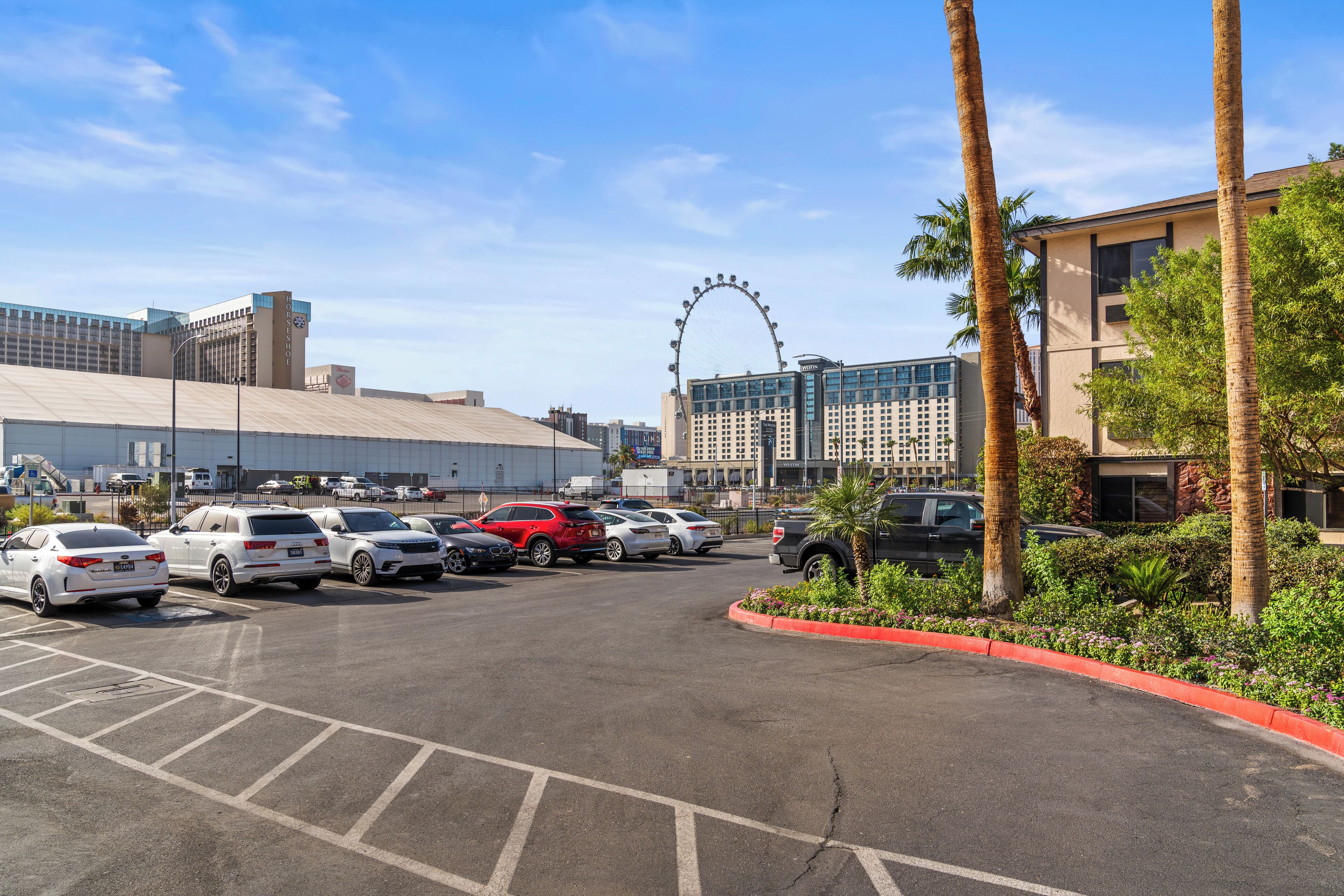 A parking lot with several cars, a Ferris wheel in the background, and buildings with palm trees.