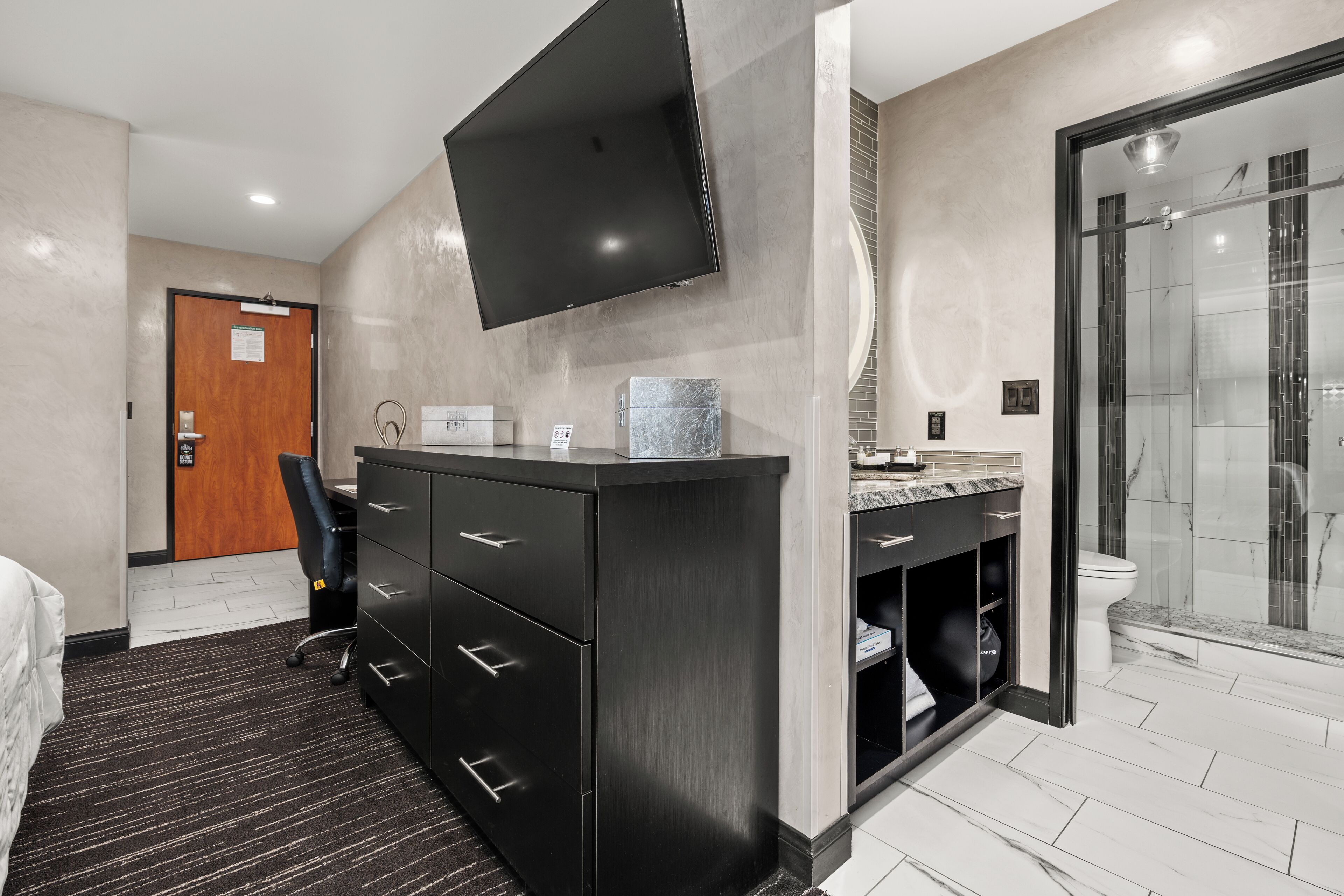 Habitación de hotel moderna con un televisor de pantalla plana grande montado en la pared, un baño independiente con ducha de vidrio y un escritorio con silla.