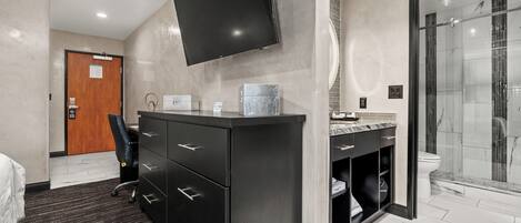 Habitación de hotel moderna con un televisor de pantalla plana grande montado en la pared, un baño independiente con ducha de vidrio y un escritorio con silla.