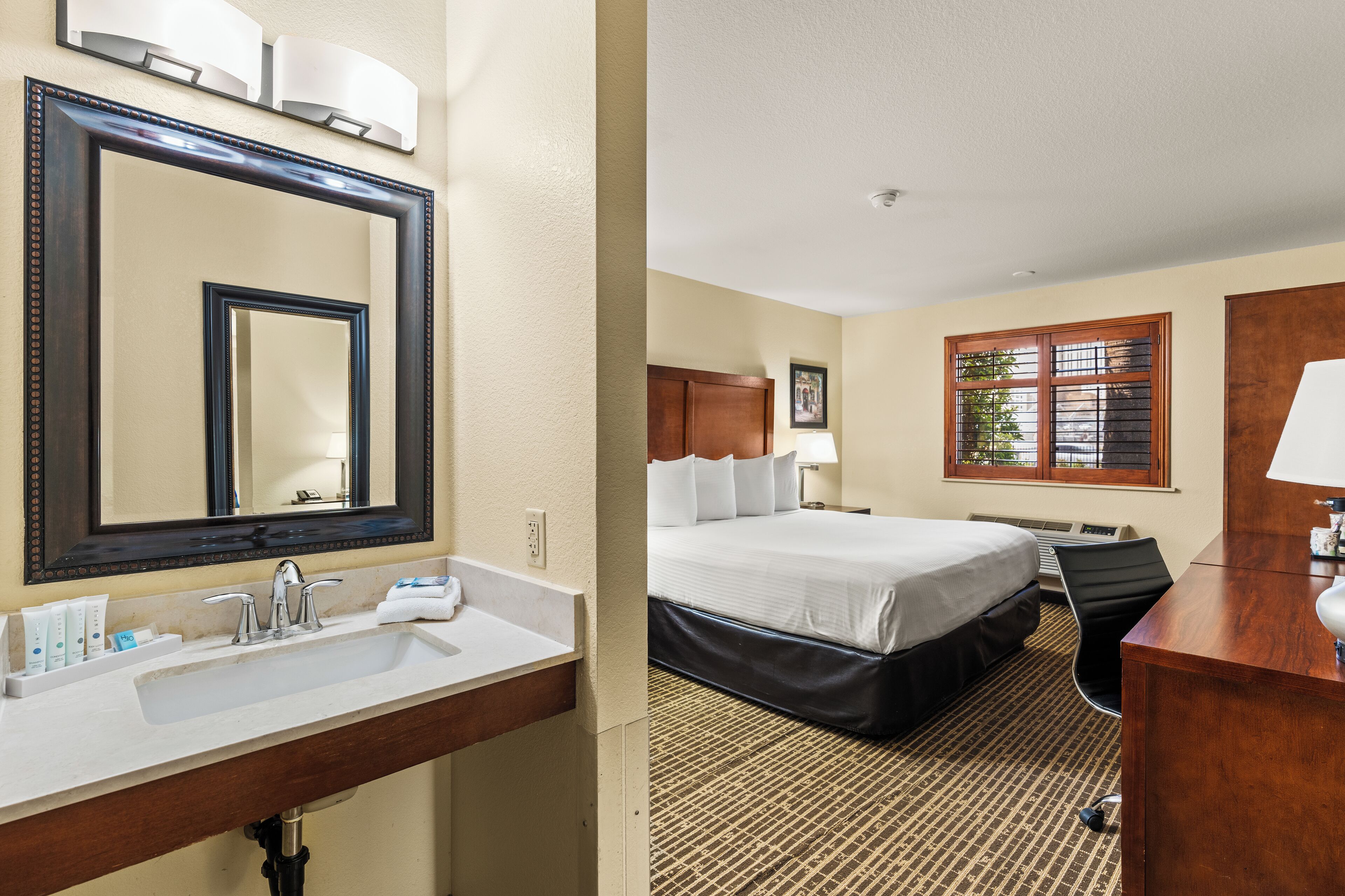 A hotel room with a large mirror, a bed, a desk, and a window with blinds.