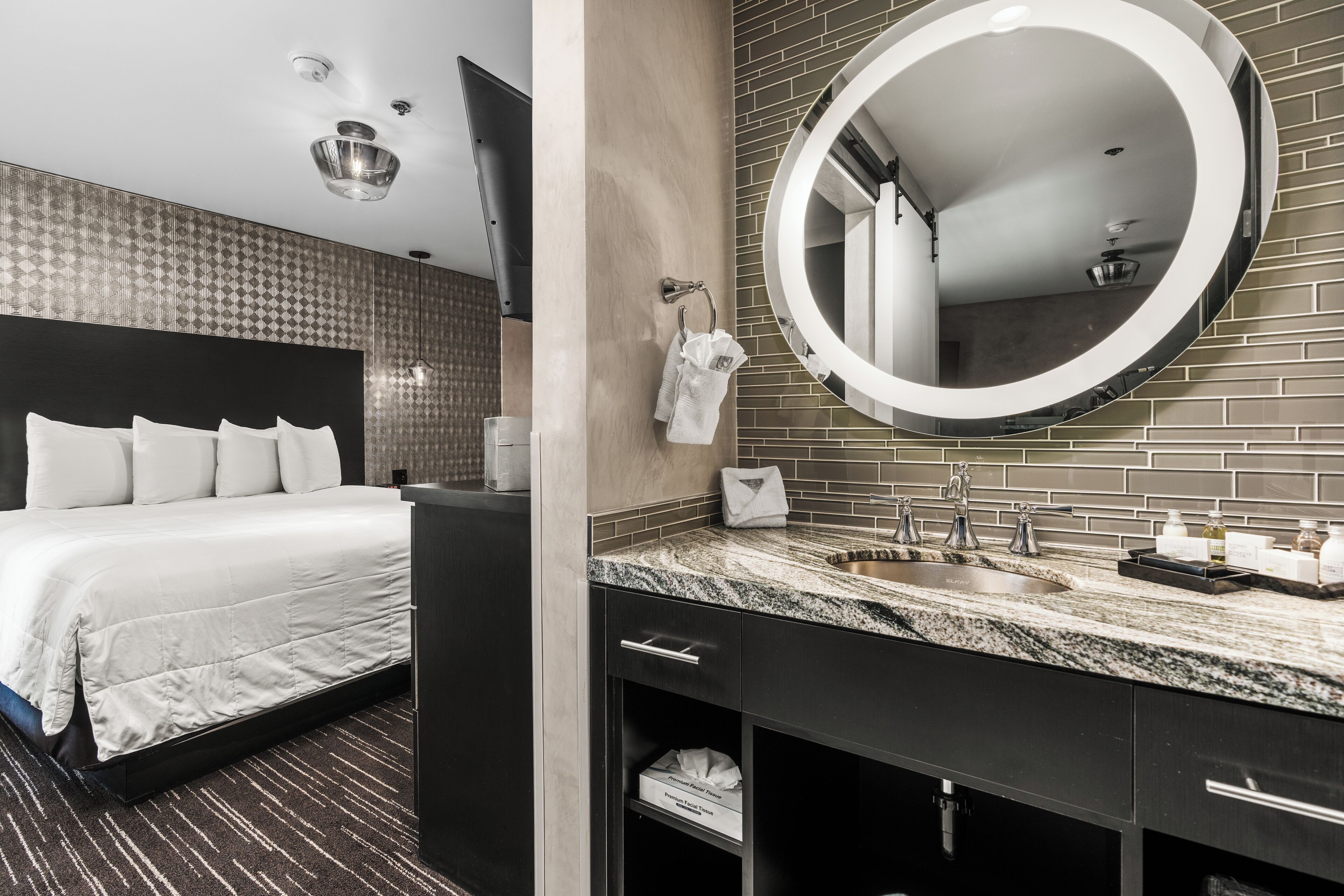 Een moderne hotelkamer met een groot bed, een ronde spiegel, een granieten aanrechtblad en een donkergrijze kledingkast.