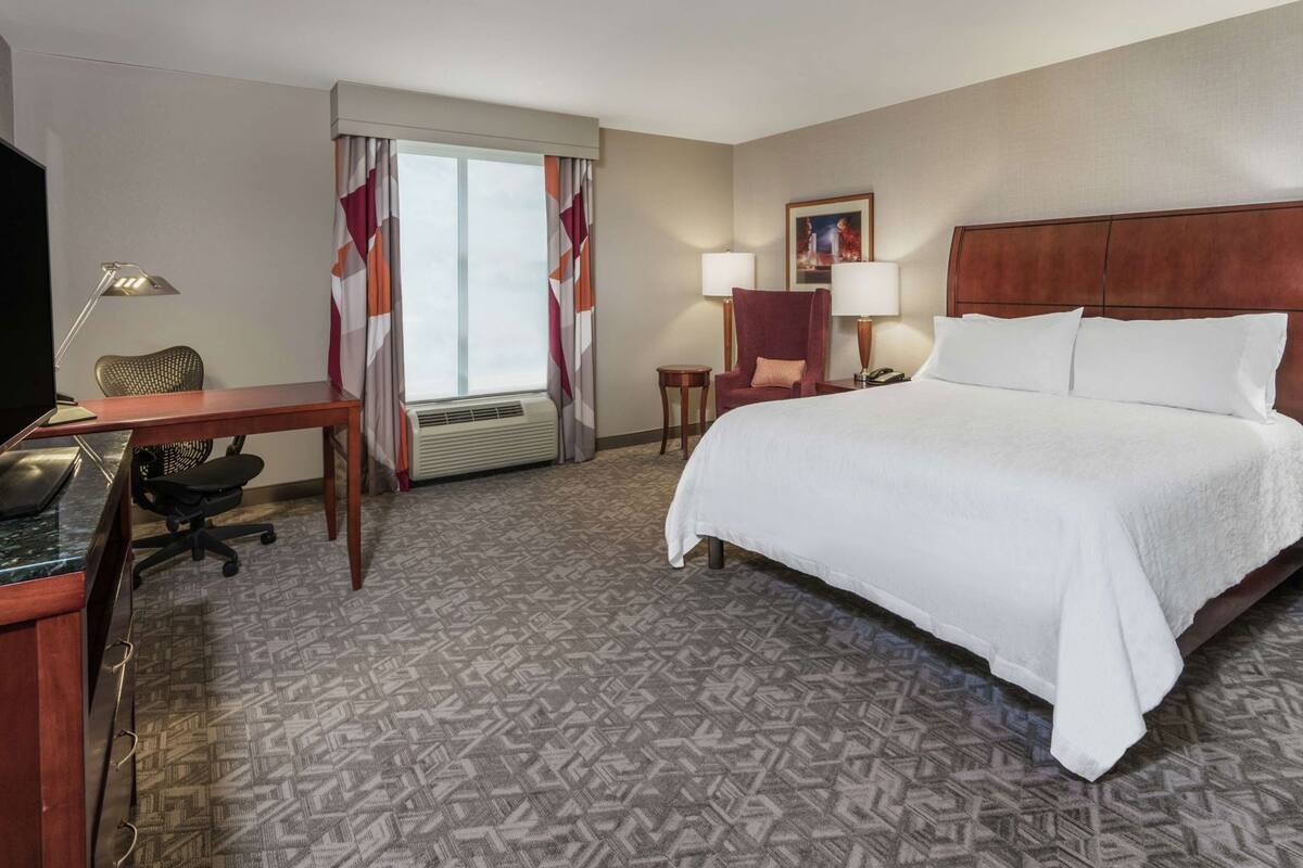A hotel room with a bed, desk, chair, lamp, and air conditioning unit.