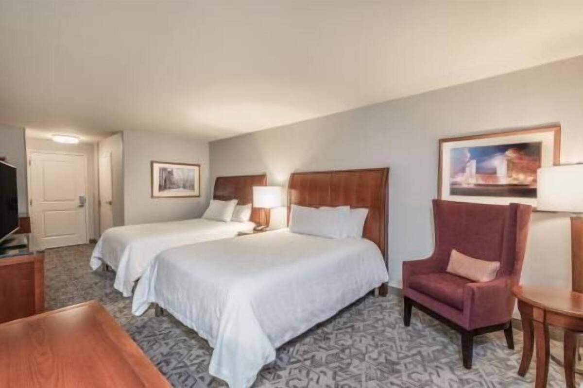 A hotel room with two beds, a desk, a chair, and a TV.