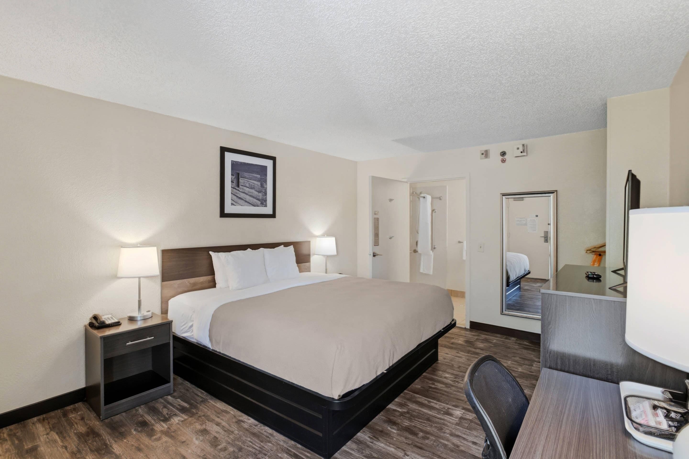 A hotel room with a bed, bedside table with a lamp, a desk with a chair, a mirror, and a door leading to another room.