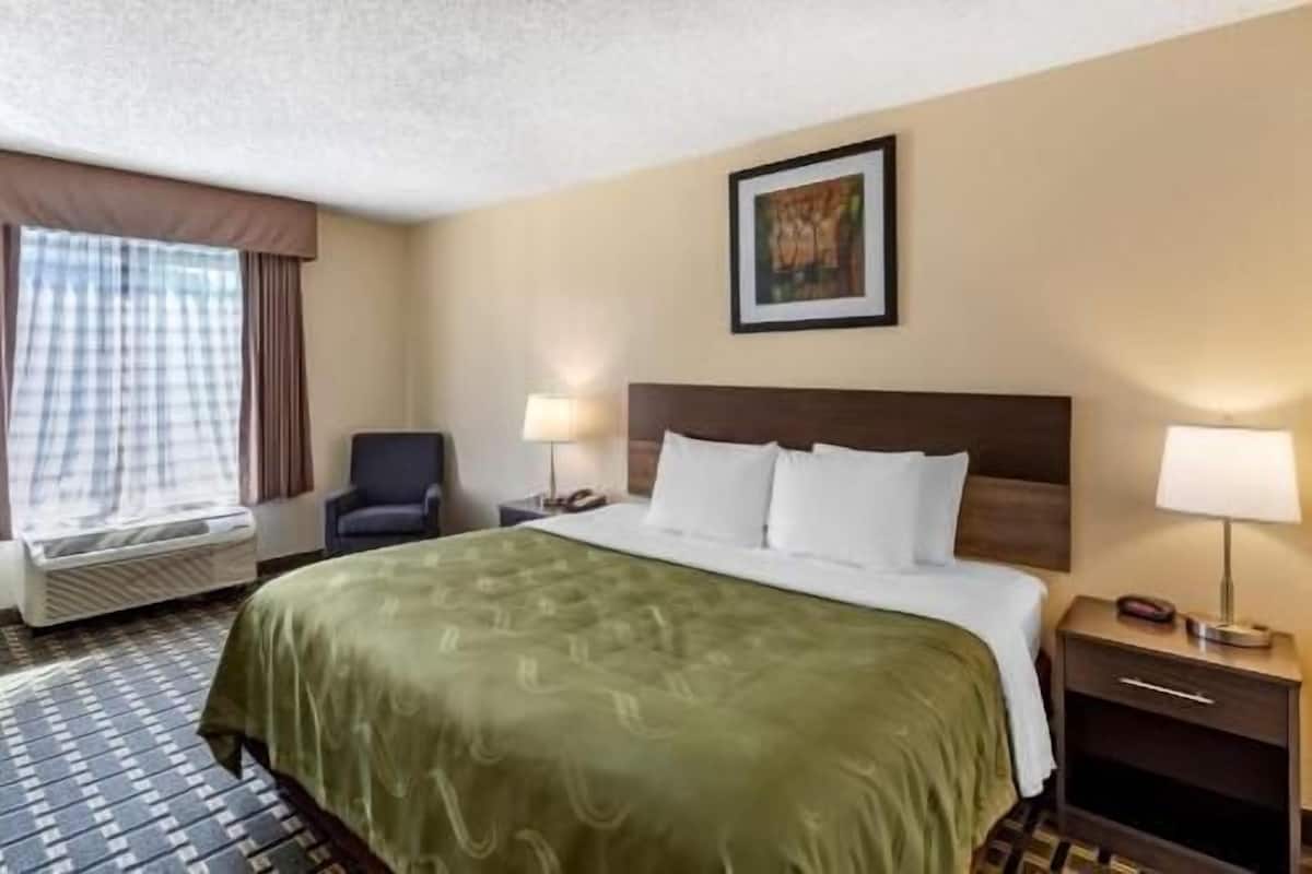 Quarto de hotel com cama, duas luminárias de cabeceira, uma cadeira, janela com cortinas e um quadro na parede.