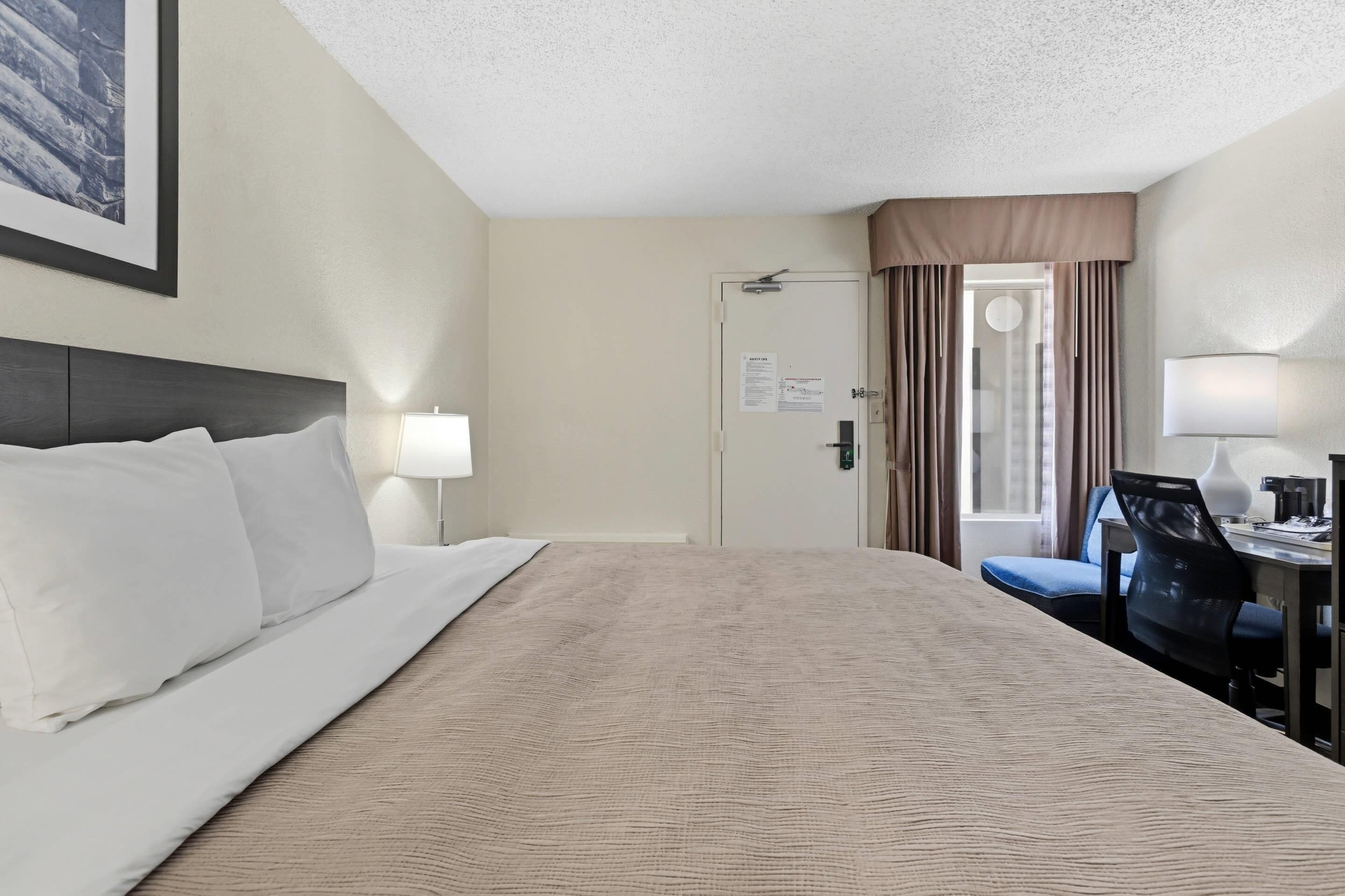 A hotel room with a bed, a desk, a chair, a lamp, and a door with curtains.