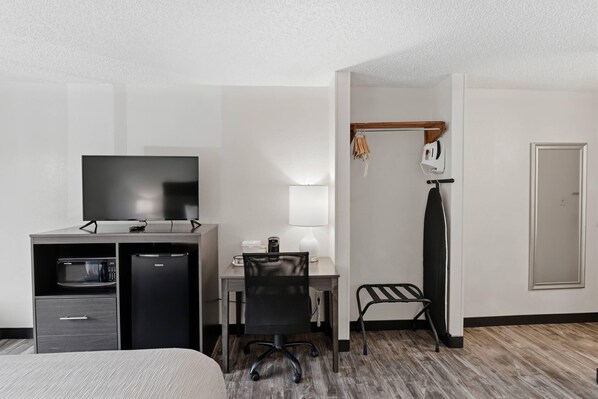 A hotel room with a bed, a desk with a chair, a television, a microwave, a refrigerator, a lamp, a coat rack, and a door.