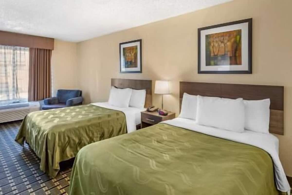 Quarto de hotel com duas camas, uma cadeira, mesa de cabeceira e obras de arte nas paredes.