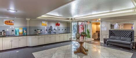 A spacious hotel lobby with a central round table, a sofa, and a kitchenette area.