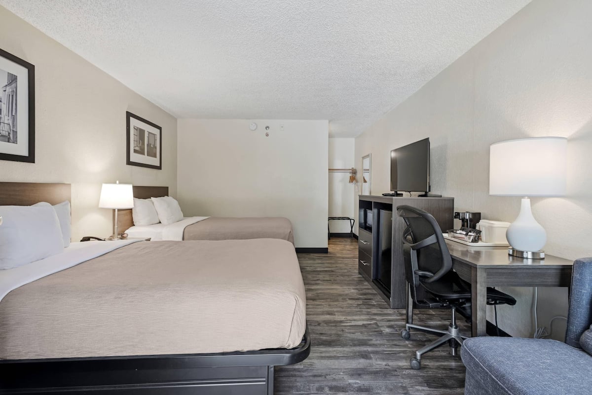 Quarto de hotel com duas camas, uma escrivaninha, uma cadeira, uma televisão e um abajur.