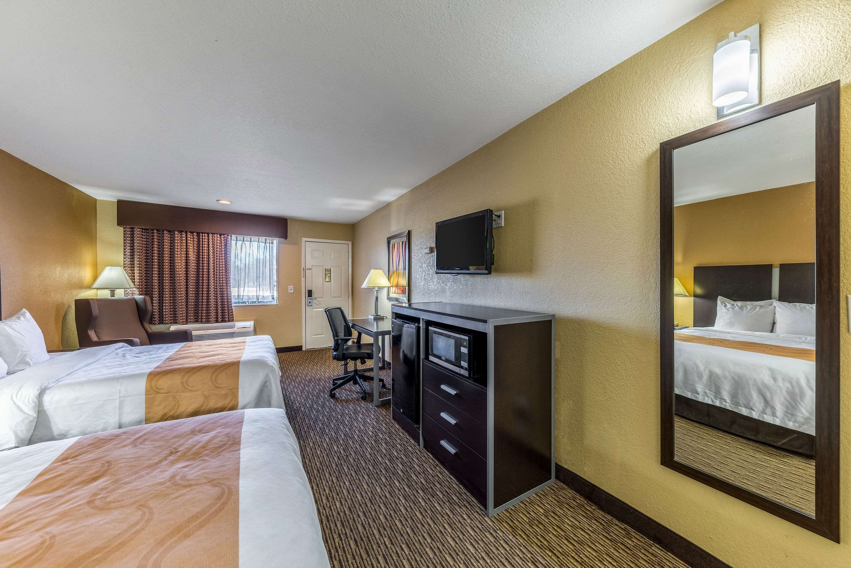 Una habitación de hotel con dos camas, un escritorio con computadora, un televisor y un espejo grande.