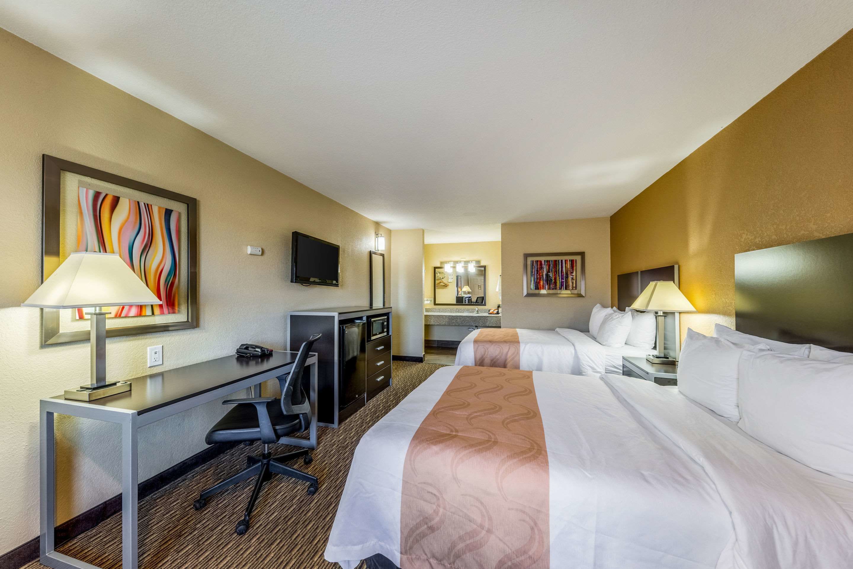 A hotel room with two beds, a desk with a chair, a television, and a bathroom with a mirror and sink.
