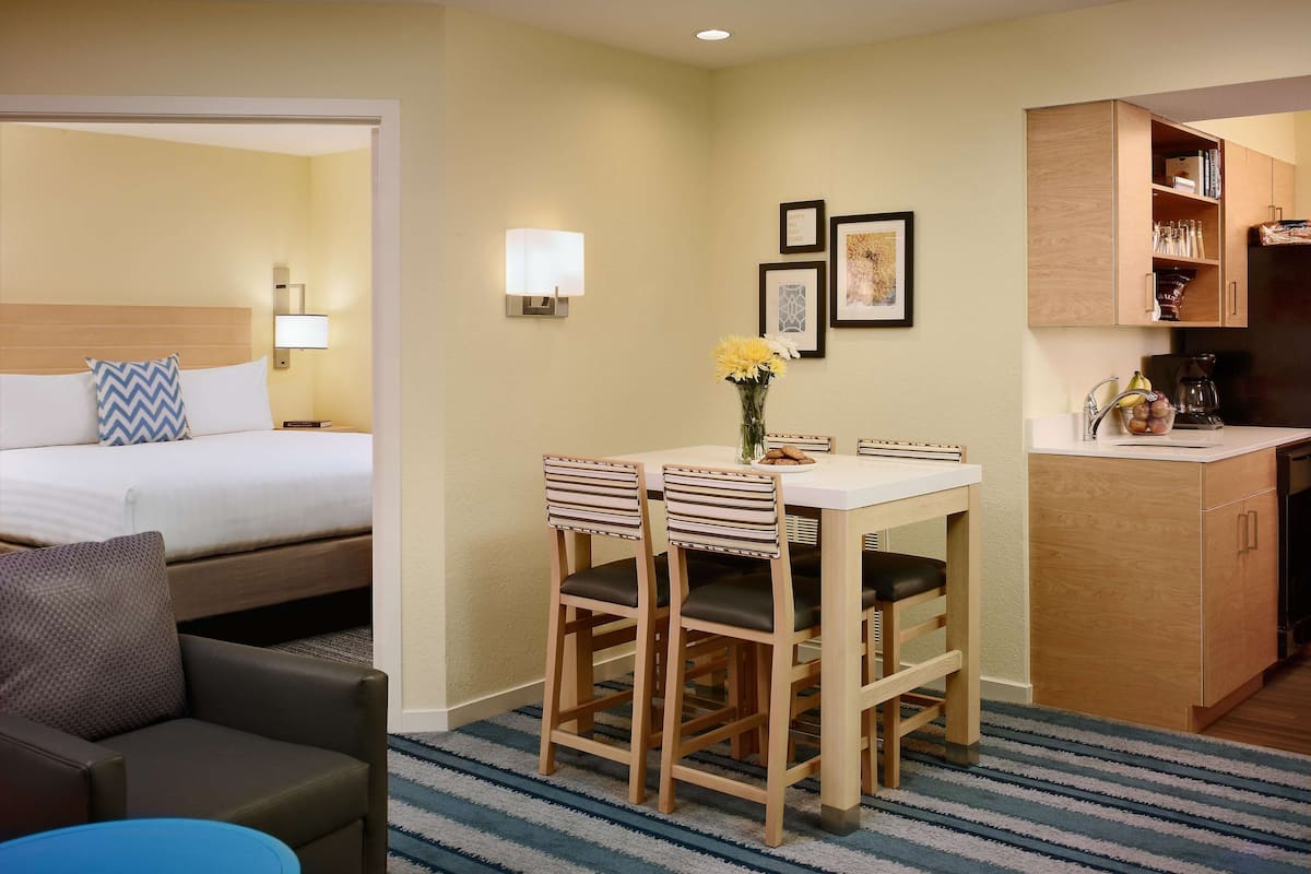 Habitación de hotel con cama, escritorio con sillas, kitchenette pequeño y alfombra azul a rayas.
