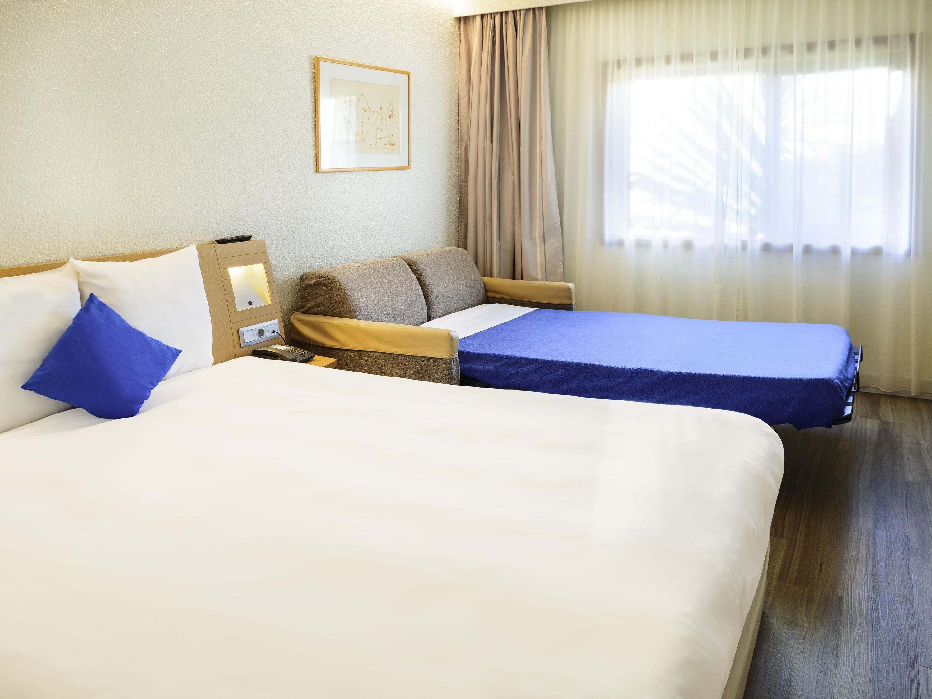 A hotel room with two beds, a bedside table with a lamp, a framed picture on the wall, and a window with curtains.
