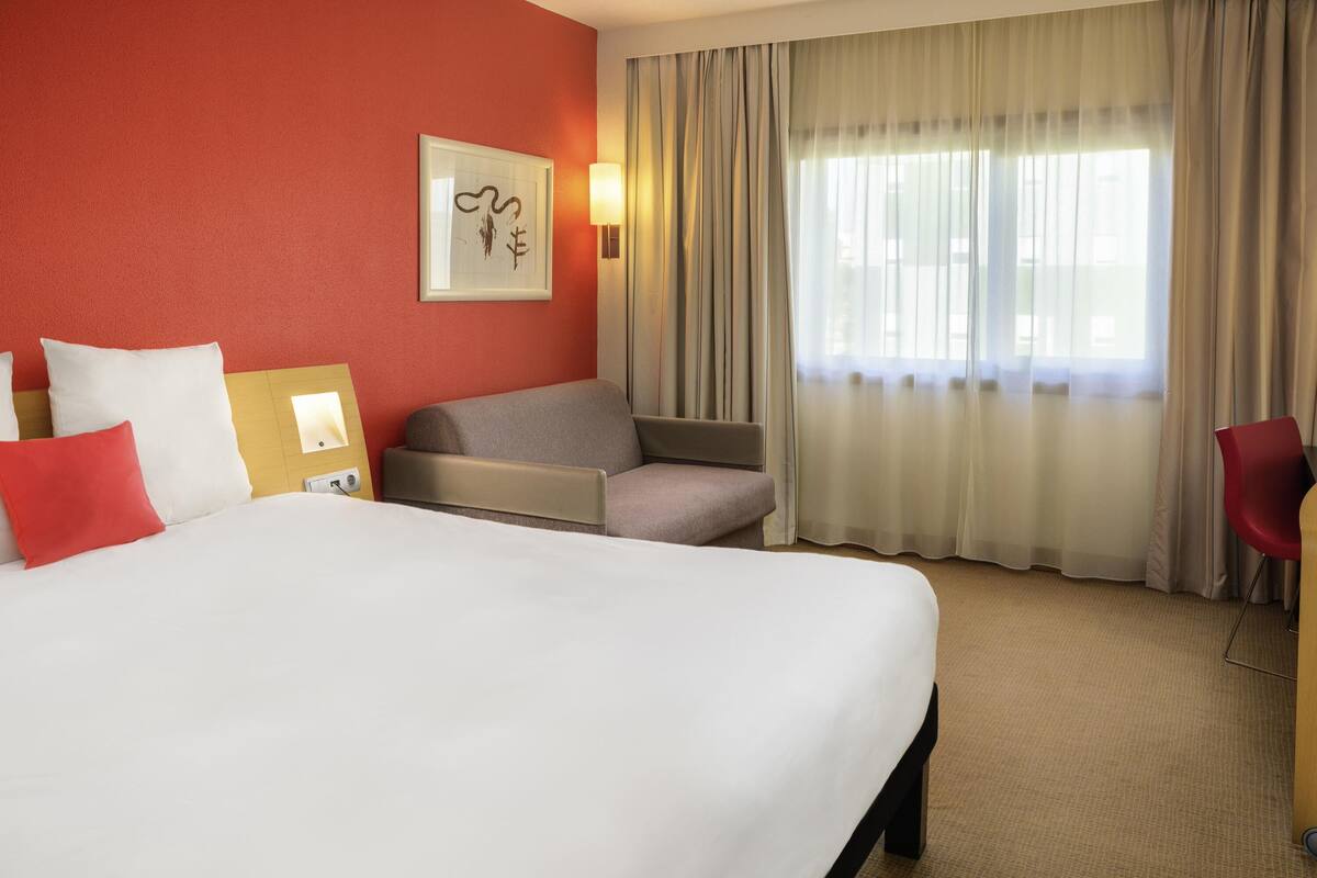 Habitación de hotel con una cama grande, un sofá, una silla y un escritorio. Hay una ventana con cortinas, una lámpara de pared y una obra de arte enmarcada.