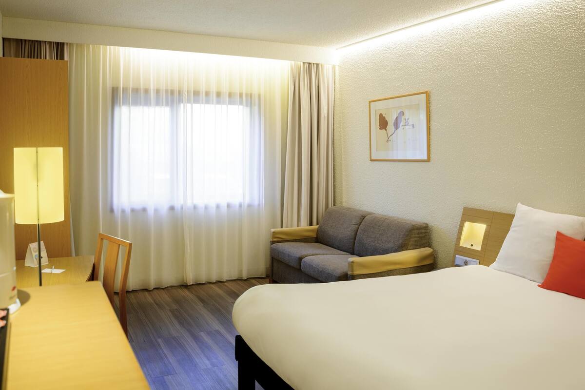 Una habitación de hotel con cama, sofá, escritorio con lámpara y ventana con cortinas.