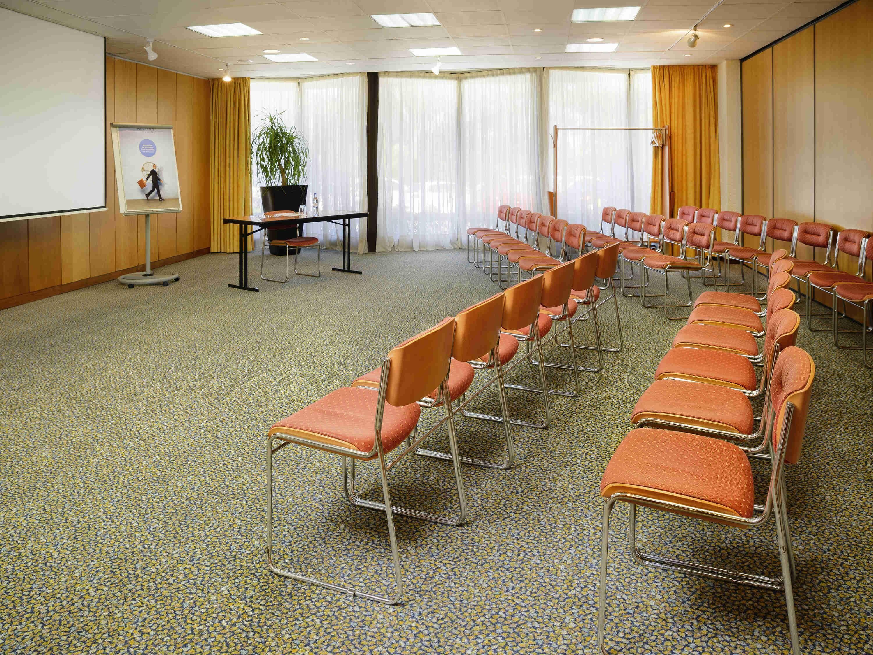 A conference room with rows of orange chairs, a projector screen, and a large window with curtains.