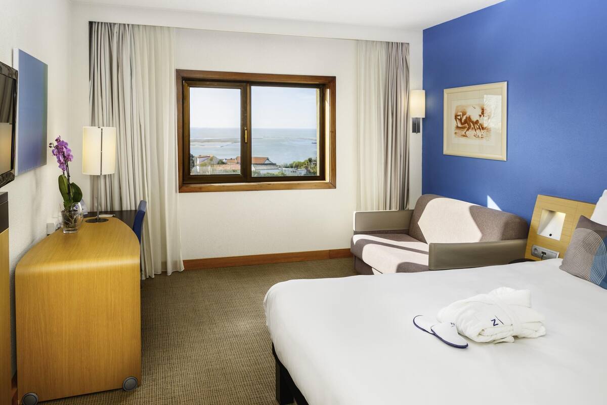 Habitación de hotel con cama, escritorio, silla, lámpara, ventana con vista al mar y un cuadro en la pared.