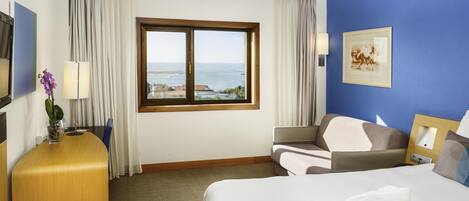 A hotel room with a bed, a desk, a chair, a lamp, a window with a view of the sea, and a painting on the wall.