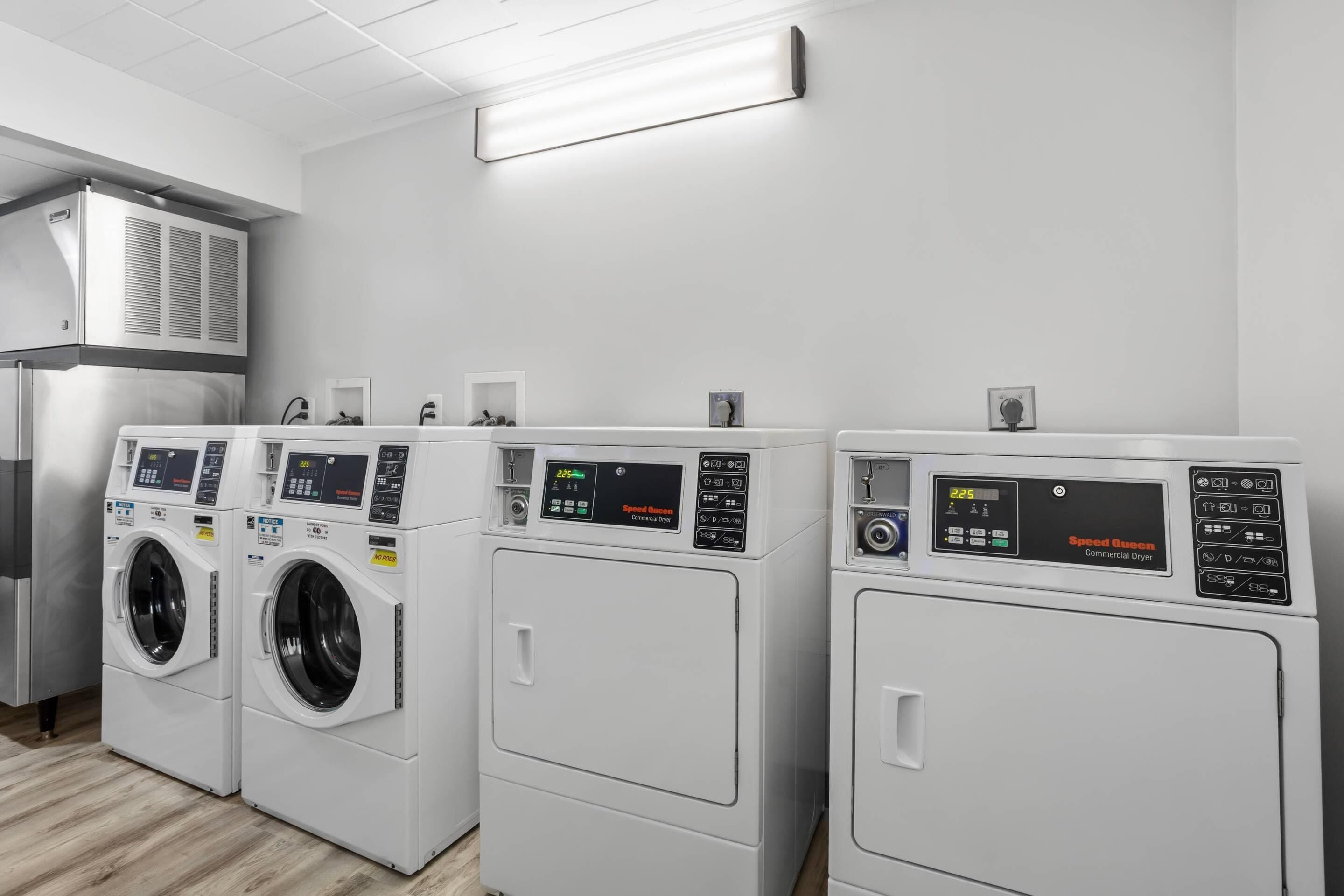 A laundry facility with three Speed Queen commercial dryers, a stainless steel refrigerator, and a white wall with a ceiling light fixture.