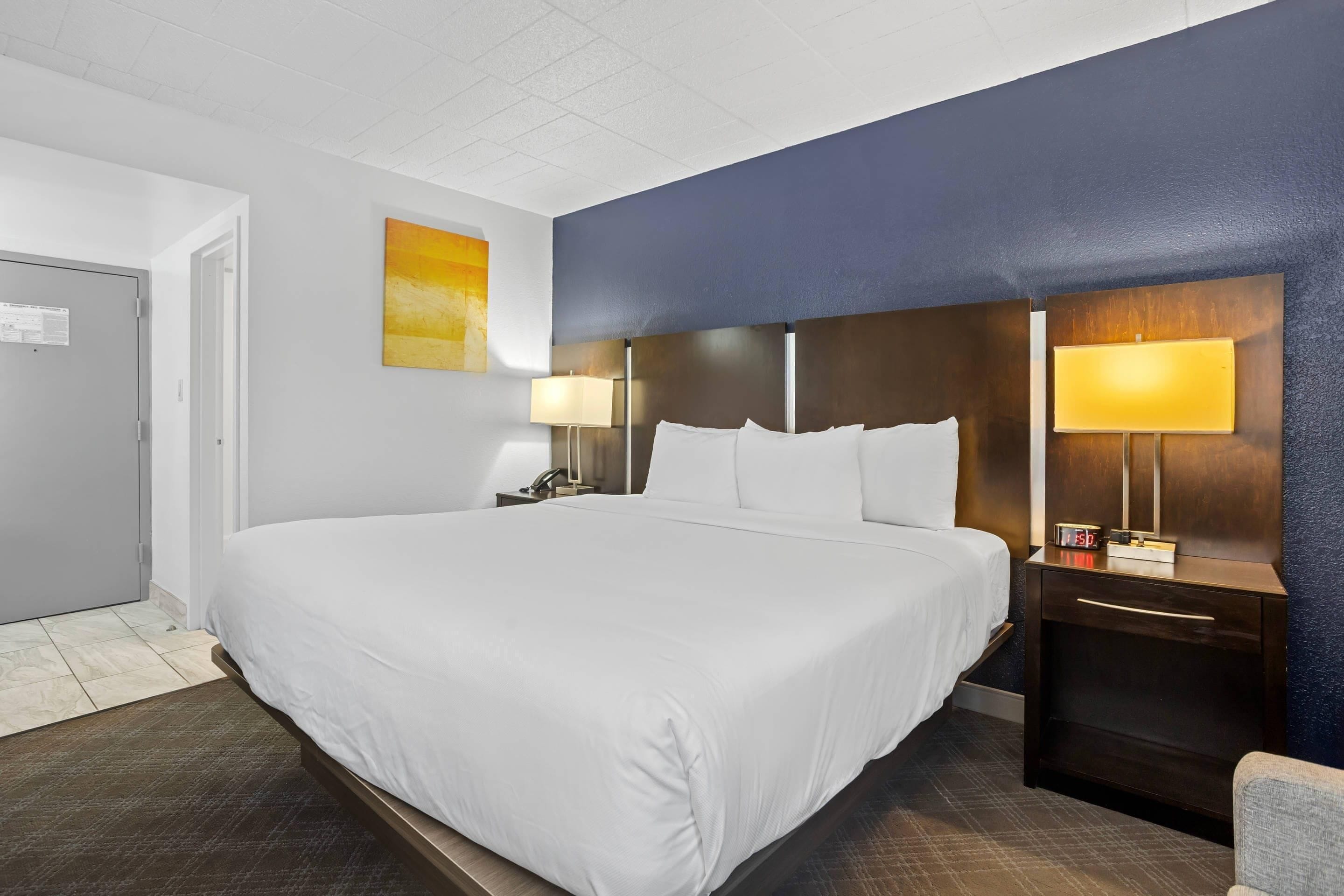 A hotel room with a large bed, a nightstand with a lamp, a wall clock, and a door.