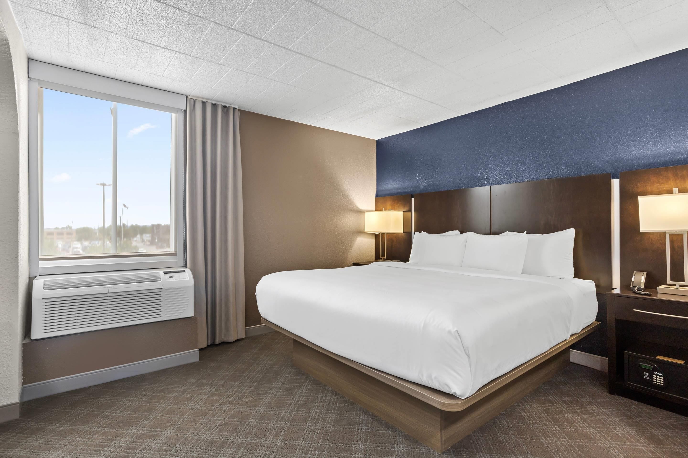 A hotel room with a large bed, a window with curtains, a wall-mounted heater, a bedside table with a lamp, and a control panel.