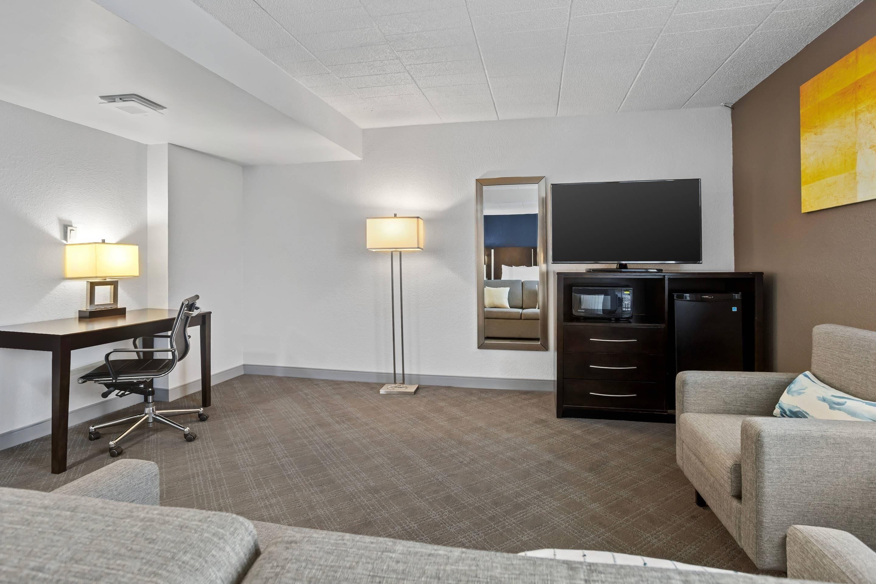 Habitación de hotel con escritorio, silla de oficina, sofá, lámpara de pie, televisor y minibar.