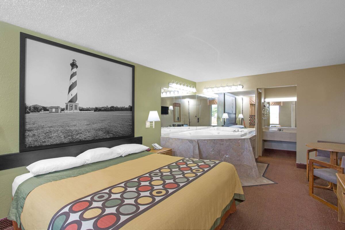 A hotel room with a bed, a large painting of a lighthouse, a bathroom with a sink and mirror, and a chair.