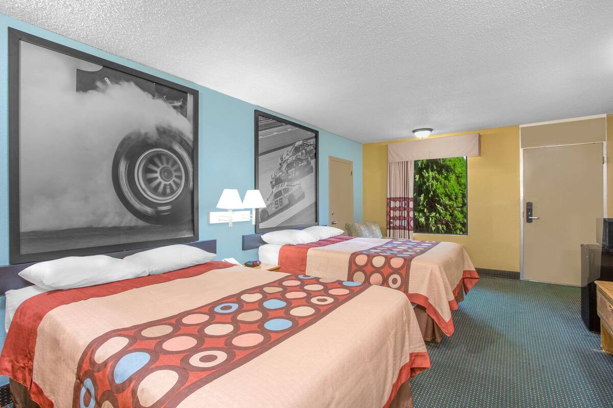 A hotel room with two beds, a large poster of a car, a TV, and a window with a view of greenery.