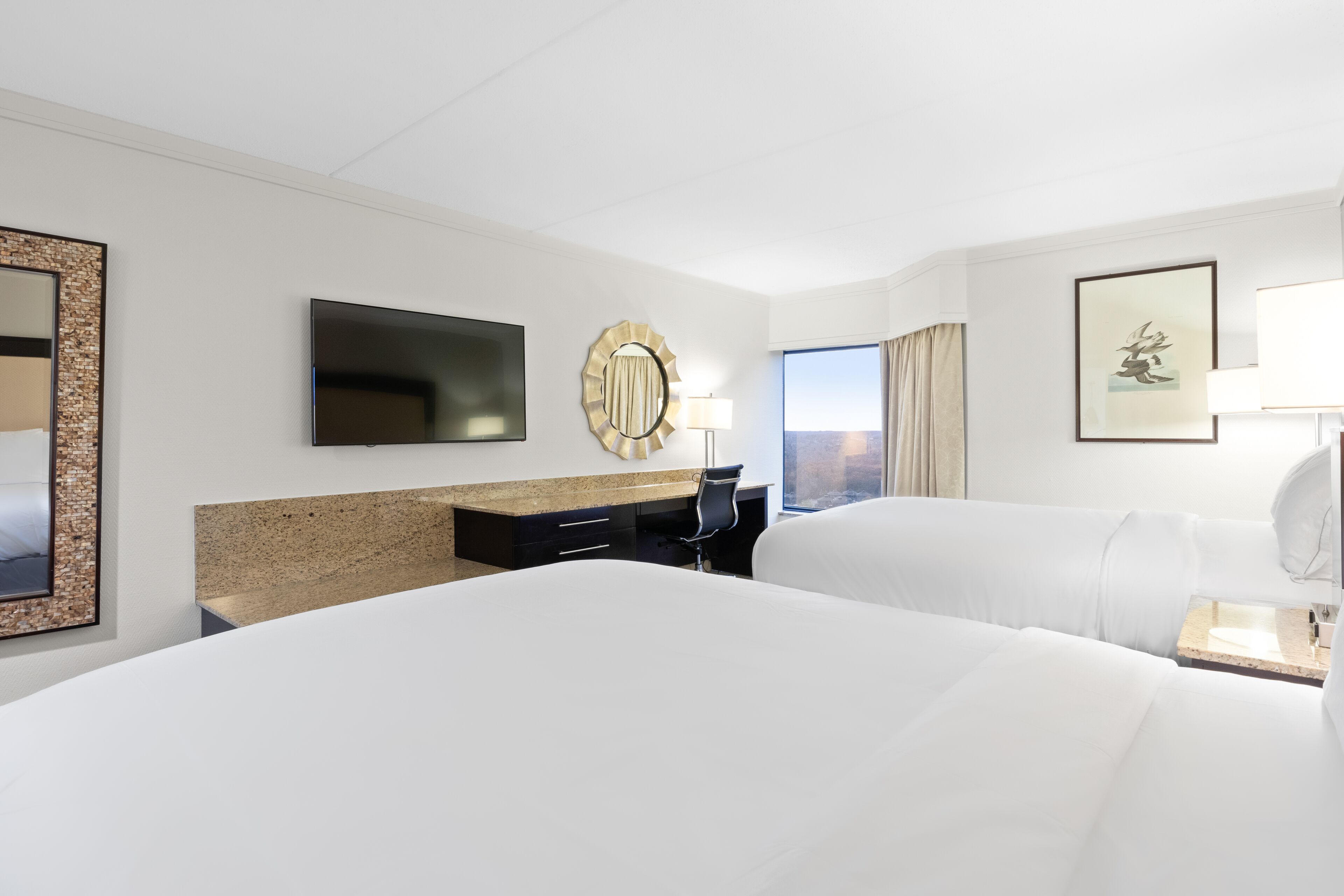 Habitación de hotel con dos camas, un televisor de pantalla plana, un escritorio con una silla, un espejo redondo y una ventana con vistas.