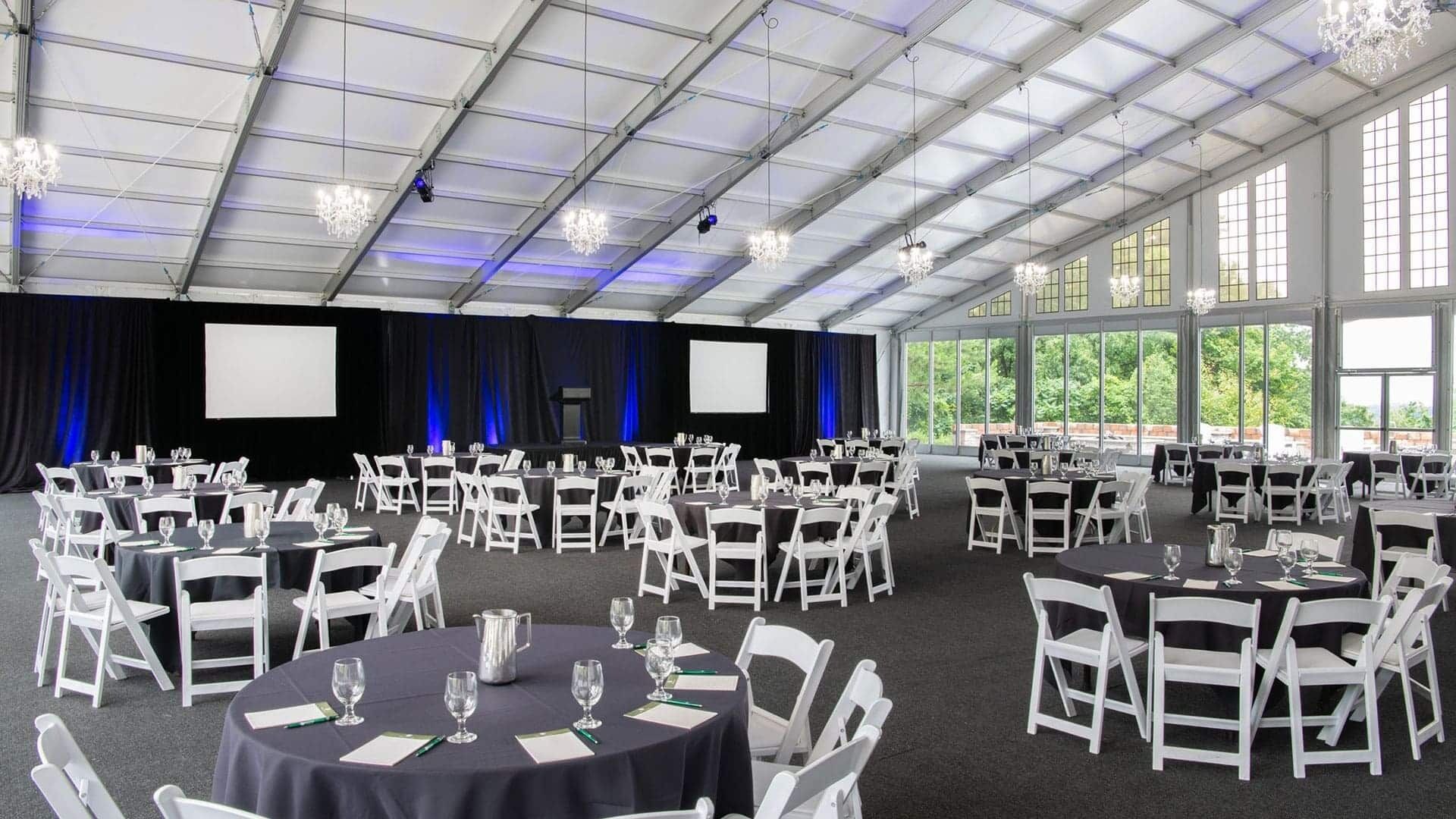 A large indoor event space with multiple round tables, white chairs, and a stage with a screen.