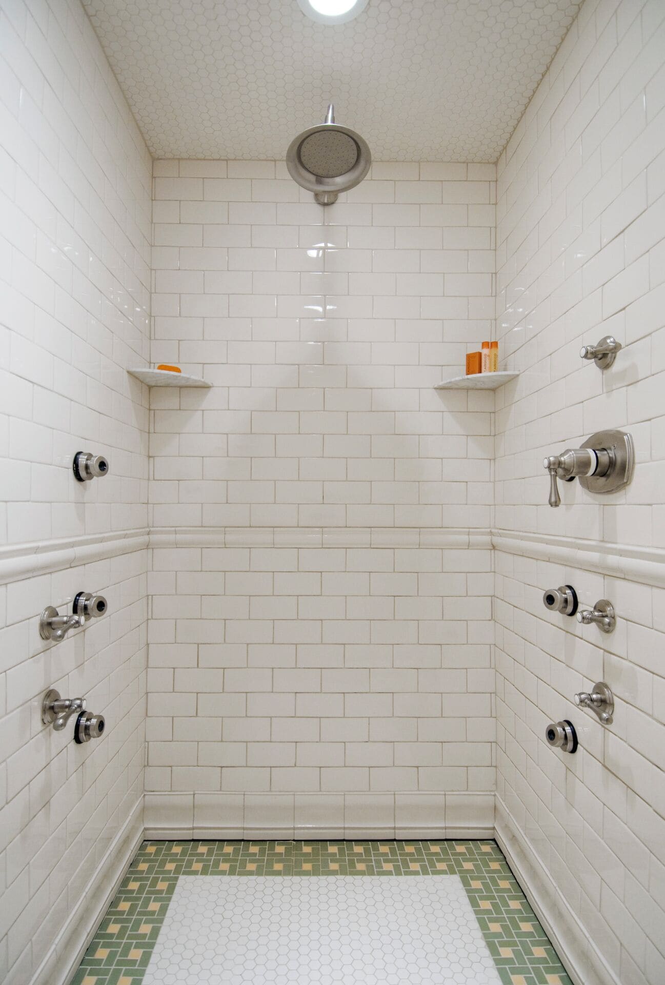 Une petite douche carrelée avec un plafond blanc, un sol à motifs verts et blancs, et une seule étagère où sont rangés les articles de toilette.