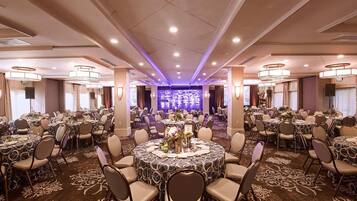 A large banquet hall with round tables, chairs, and a stage with a blue backdrop.