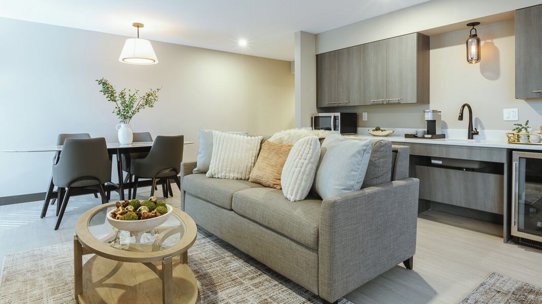 A modern living room with a sofa, a dining table with chairs, a kitchen area with cabinets and appliances, and a small round table with a bowl of fruit.