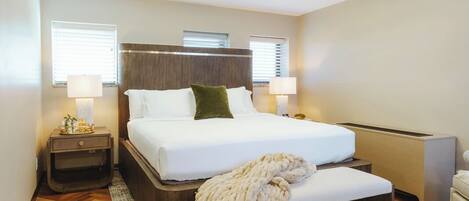 A modern hotel room with a large bed, bedside lamps, a wooden headboard, and a small bedside table.