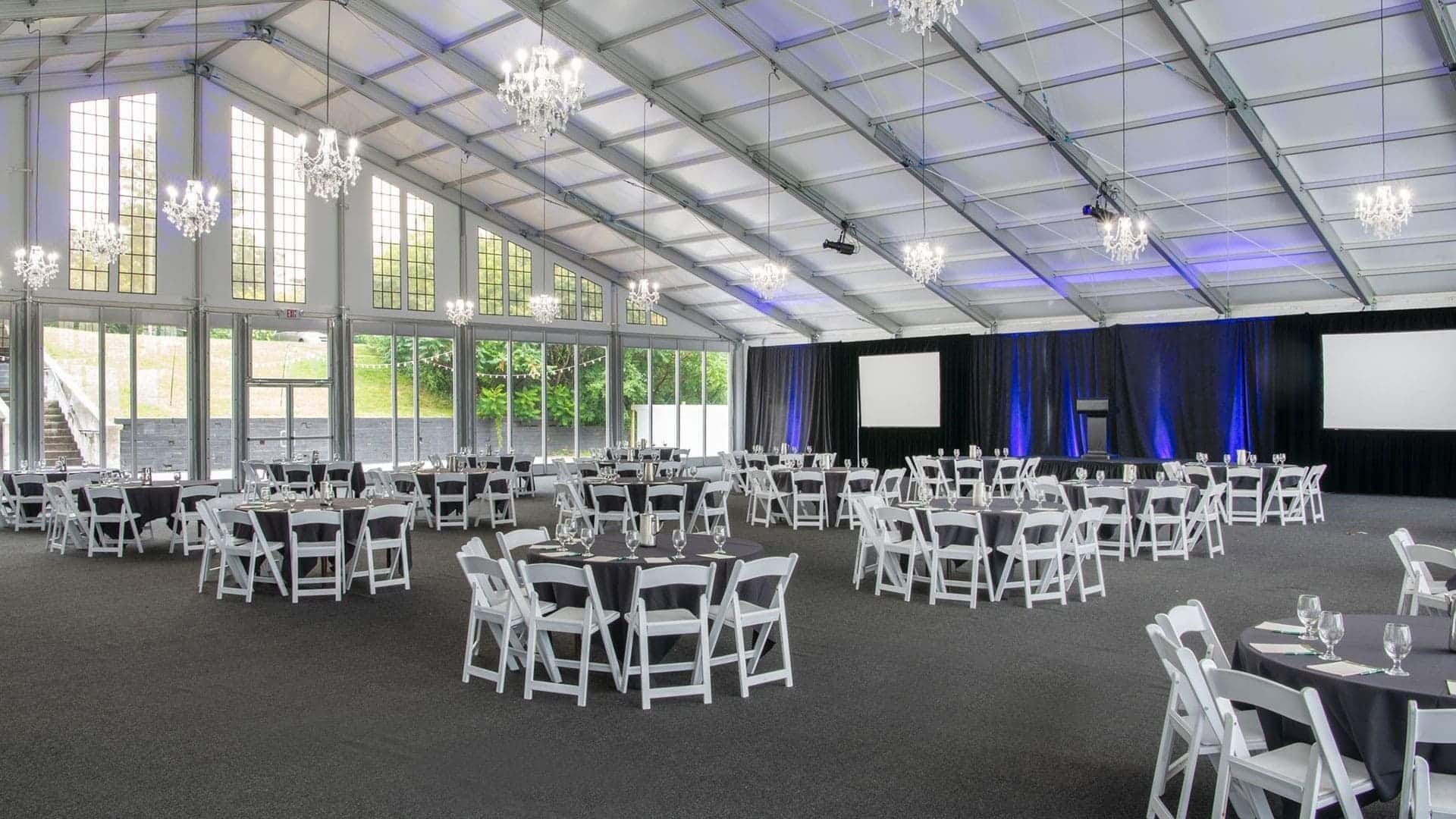A large indoor event space with rows of round tables and white chairs, a stage with a screen, and chandeliers.