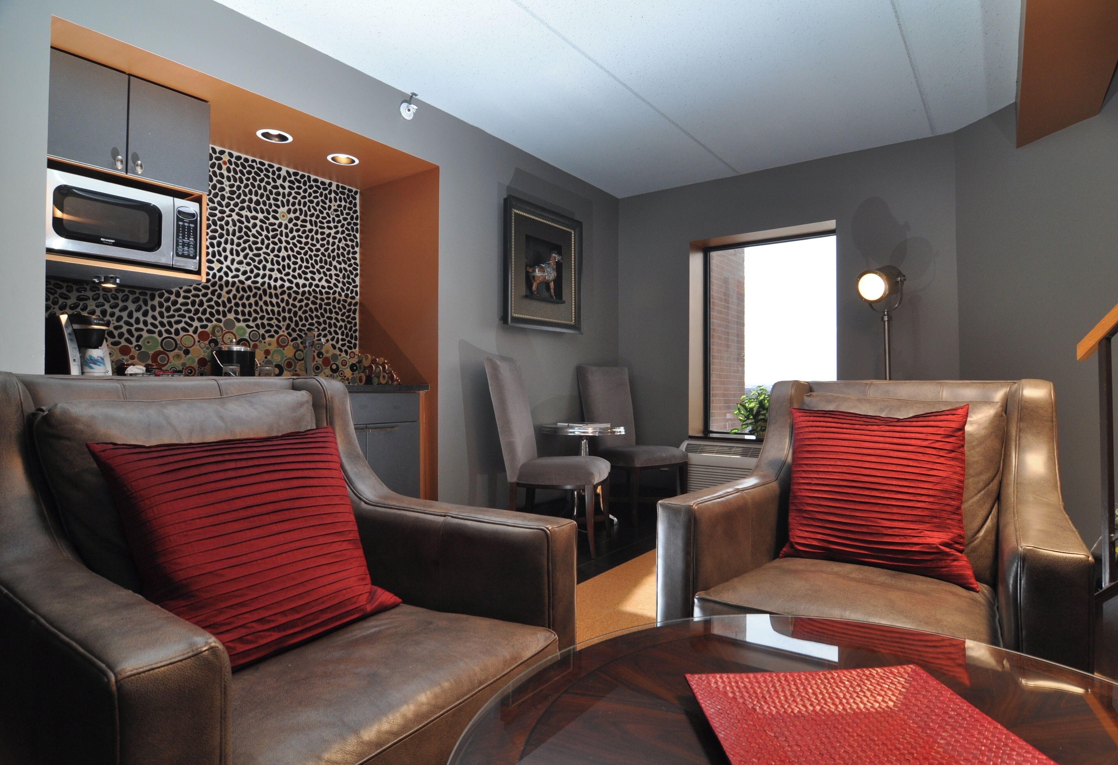 A modern living room with leather armchairs, a glass coffee table, a kitchen area with a microwave, and a framed picture on the wall.